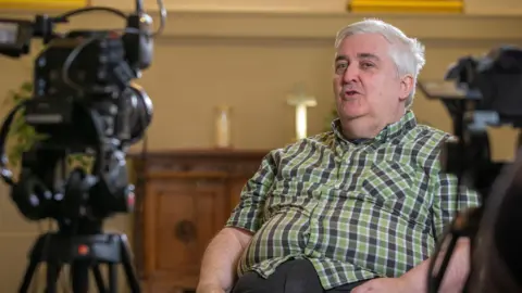 Daniela Cotognini A man sits on a chair in front of two cameras, speaking to someone who is not in frame.