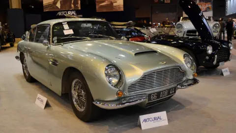 Getty Silver-grey classic Aston Martin-style coupe displayed at an indoor collector car show, viewed from the front-left angle. The car features a large chrome grille, round headlights, wire-spoke wheels, and polished chrome trim. Display signs reading “ARTCURIAL MOTORCARS” sit in front of the vehicle, while other vintage cars and exhibition displays are visible in the background under bright showroom lighting.
