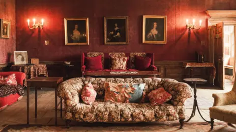 Saltwick Media /Bav Media The interior of a stately home. The walls are painted red, with four portrait paintings. There are several sofas, with flowery printing, as well as several small wooden tables.