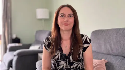 BBC News Sarah Todd sitting on a grey sofa, wearing a black top with white leaf print. She wears her dark brown hair over her shoulders and has a silver necklace on. She is smiling at the camera. In the background, you can see the sofas and a white lamp behind her, which is out of focus. 