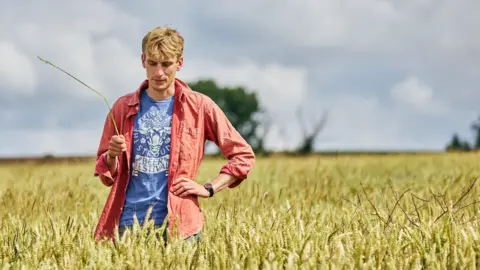 Cirencester History Festival Charlie Cooper stands in a field of tall grass, holding a blade of it, as he looks down. He is wearing a blue T-shirt with a white Stroud Beer Festival logo on and a dusky reddirt over y pink shthe top. There are trees in the hedgerow behind.