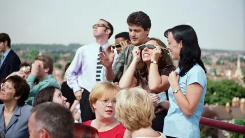 Getty Images A group of people sitting and standing while waiting for a solar eclipse to take place. Some people are wearing solar eclipse glasses.