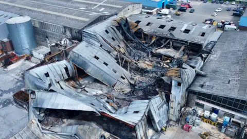 Ant Saddington/BBC The scene of the fire from the air, with large sections of twisted and charred metal with small window holes visible. There is a large metal tank to the left.