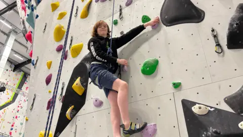 Sam wearing a harness on a rope whilst climbing a wall with different coloured holds. He is facing the camera and smiling.