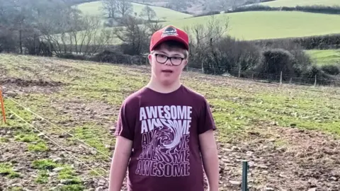 A boy is standing in a field. He is wearing a red cap and a T-shirt that says "awesome" several times, with a swirl design in the words.