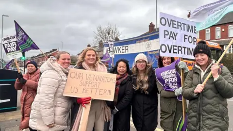 Woman gathered wearing coats and holding placards outside of a hospital setting.