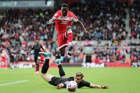 Tom Banks/MFC A Middlesbrough player in all red chases a ball as he jumps over another player all in black lying on the ground.