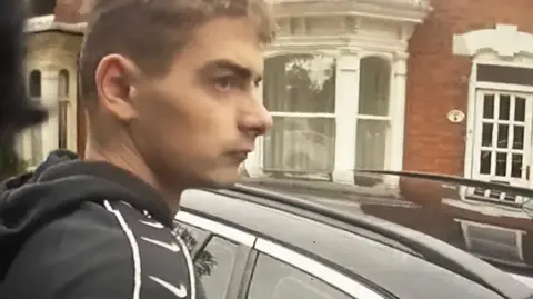 Lincolnshire Police A young man wearing a black tracksuit is captured on police body worn cameras as he is arrested. He stands next to a black car. 
