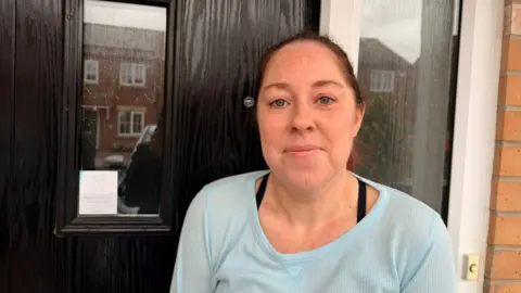 A woman stands in the doorway of her home. She has brown hair, tied back, and is wearing a light blue top. The door is black.