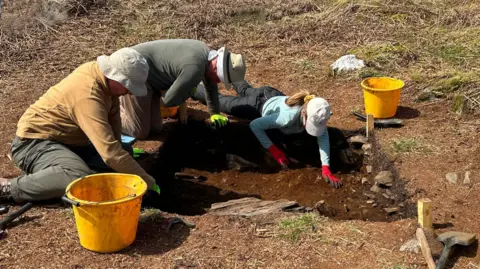 Antiquity Publications Ltd Three people wearing a range of clothing digging in a hole during the excavation. There are yellow buckets beside them.