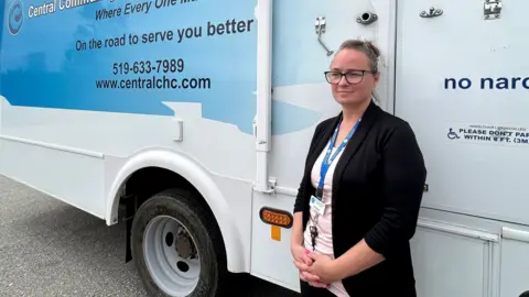 O Canadian Press Catalina Friesen, um trabalhador de apoio pessoal e um ligação de baixa língua alemã, fica em frente a um ônibus equipado como uma clínica móvel, em St. Thomas, Ontário. Ela tem um leve sorriso no rosto e está usando um blazer escuro e uma camiseta branca