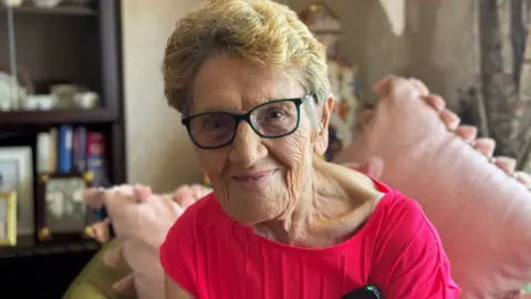 Mary Brown is an older woman with short blonde hair. She is smiling at the camera and is wearing dark framed glasses. Behind her are two large pale pink cushions with tassels and a dark bookcase with books, photo frames and trinkets on it. 