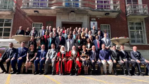 A large group of councillors in suits and other smart clothes sit in rows for the camera outside the council house in Swindon. In the centre on the front row are two ceremonial councillors in their red robes with black trim