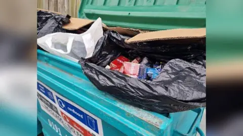 Black bin bags containing cigarettes and vapes sit on rubbish in a large, blue commercial wheelie bin.