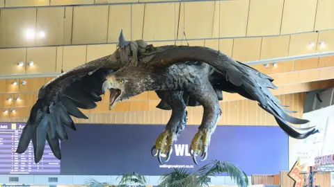 Side view of the giant eagle hanging from the ceiling, with Gandalf riding on its back. Behind the sculpture is a large purple screen showing flight arrival and departure times.