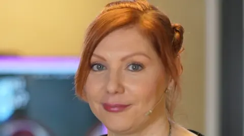 Jamie Niblock/BBC Hannah Aria smiles at the camera. She hair red hair that has been tied up. She also has a microphone piece resting against her cheek. 