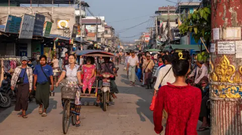 Jonathan Head/ BBC A busy street in a market in Yangon. People are seen walking down it, alongside rickshaws carrying people. 