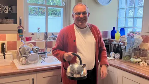 Andrew in a white T-shirt and orange cardigan standing holding a sinver kettle in his kitchen