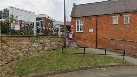 Google A red brick building with a black iron fence and multi coloured bunting draped over the fence. 