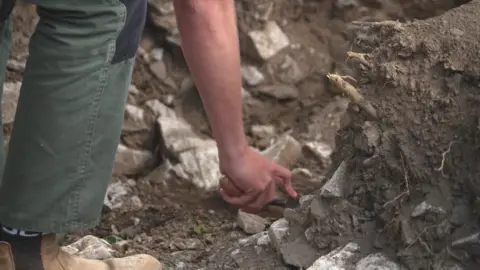 An archaeologist is uncovering part of a stone wall in a trench.
