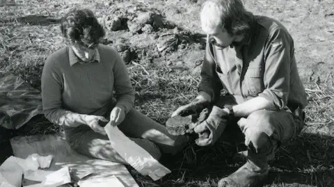 Shropshire Museums A black and white photo of a man and a woman. The woman is sitting on a wooden board on grassy and muddy ground, while the man crouches down beside her. The woman is about to open a white plastic bag. The man has thick gloves on, and is holding a piece of bone