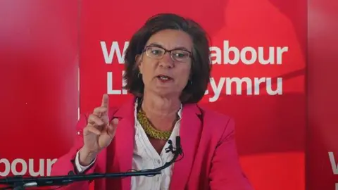 PA Media Eluned Morgan speaking in front of a red Welsh Labour banner, wearing a pink jacket. She has a white shirt underneath with a gold chunky necklace on. She has purple framed glasses and dark brown shoulder-length hair.
