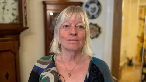 A head and shoulders photograph of Chris Suich, a woman with shoulder-length blonde hair who is wearing a green and blue top and standing in a room with a dark-brown grandfather clock and a matching cabinet behind her.