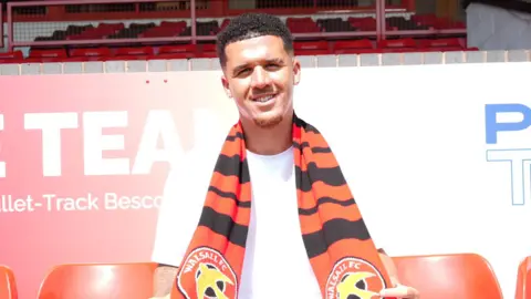 Rico Browne is sitting in the stands at Pallet-Track Stadium with a Walsall scarf around his neck. He is wearing a white tee shirt and shorts. There are red empty seats next to him on either side.