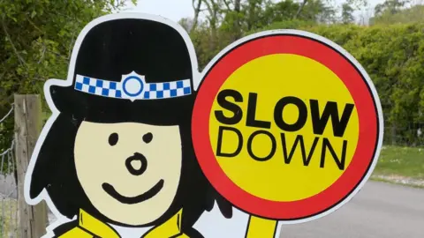 A sign outside a school with a cartoon-like depiction of a lollipop lady with the words "slow down" written on the lollipop.