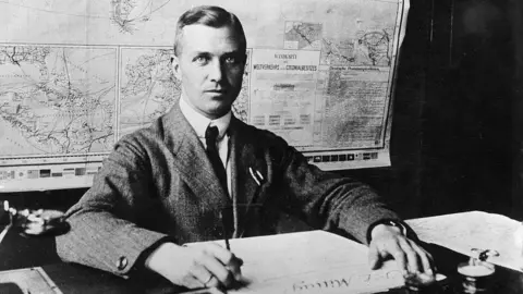 A black and white photo of a man sitting on a desk. He is holding a quill. 
