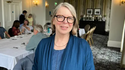 A woman with short light hair wearing a blue cardigan. She is stood in a function room with people sat at a table behind her.