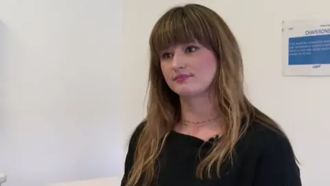 A lady with brown hair and a large fringe. She wears a black shirt.