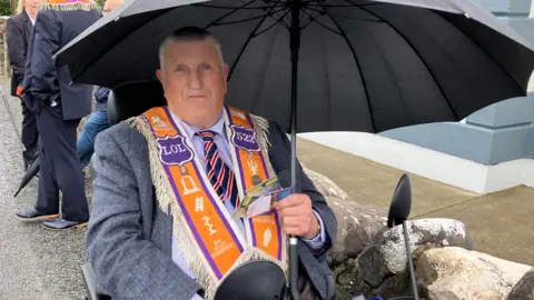 Alan Thompson in a mobility scooter and holding a black umbrella. He is wearing a grey suit, and has an orange order sash around his shoulders. He is looking directly at the camera.