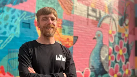 Ed Wellard stands with his arms crossed in front of a colourful pink and blue mural featuring flowers and pigeon in Withington. 