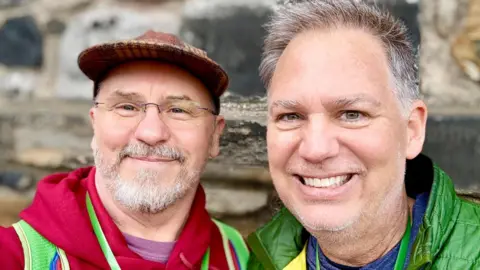 Two men are standing inside a croft house holding a sheep fleece, one of them is wearing glasses and a red hoodie and the other is dressed in a green jacket.