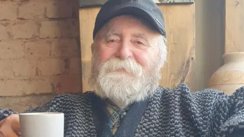 A man, Harry, pictured sat on an arm chair holding a mug. He is wearing a knitted cardigan and plaid shirt underneath. He has a full grey beard and moustache and is wearing a black cap