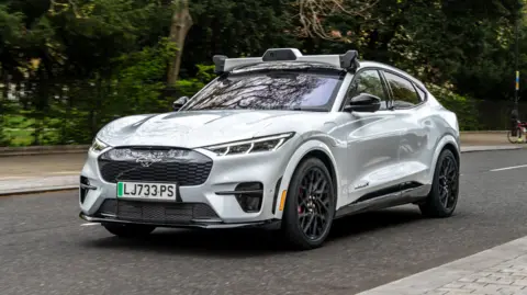 A silver electric vehicle is shown driving down a street in London. It is driving past a park, with green trees in the background.
