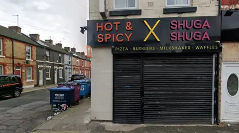 A shop has a sign saying 'Hot and Spicy', 'X' and 'Shuga Shuga' with 'pizzas - burgers - milkshake - waffles' underneath. Black shutters are down across the shop front. Industrial waste bins can be seen in a side street.