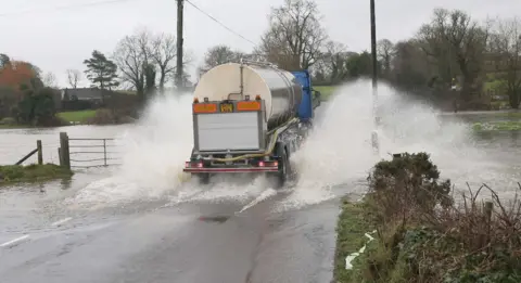 A milk tanker splashing through a flooded road. There are trees in the background.