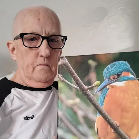 FOTO SUMINISTRADA Chris Fell, un hombre blanco calvo, lleva una camiseta blanca y gafas mientras sostiene una fotografía de un pájaro.