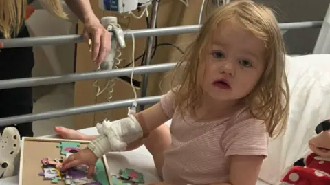 A young toddler in a hospital bed. She has a drip attached to her arm, and is playing with a jigsaw while looking into the camera while evidently unwell.