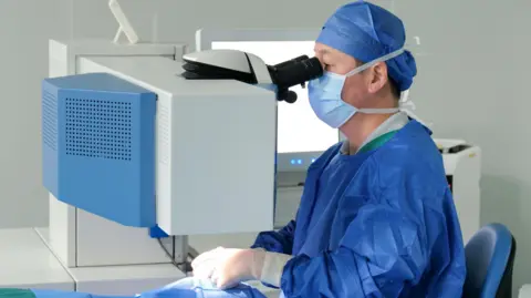Stock photo of Excimer laser surgery. A male optometrist wears all blue scrubs, and a facemask. 