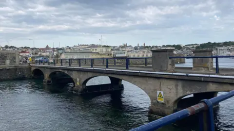 BBC The Castle Cornet Bridge in Guernsey. It is stone and has blue railings. There are some danger signs on the side of it. There is also the sea underneath it. 