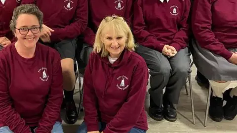 A wide shot of Judith kneeling for a full cast photo. She is smiling, and wearing a maroon hoodie. She has blonde shoulder length hair.