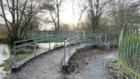 A metal bridge over a river in a wooded area, with a slope leading up to it lined with metal railings