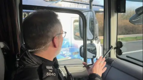 An Wiltshire Police officer in black uniform on the passenger side of the lorry cab looks through his side window towards the cab of an HGV in the adjacent lane of the M4.