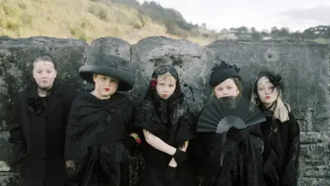 Museum Wales Five young white girls, aged approximately eight to 10, are all dressed in elaborate black dresses, veils, hats. One holds a fan. The clothing is full of lace and feathers and has a Victorian appearance overall. They stand in front of a wall which is taller than them. Behind is an out of focus grassy hill and houses in the far distance.