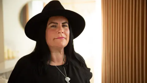 A woman wearing a black hat and a black top, standing near a window and looking at the camera