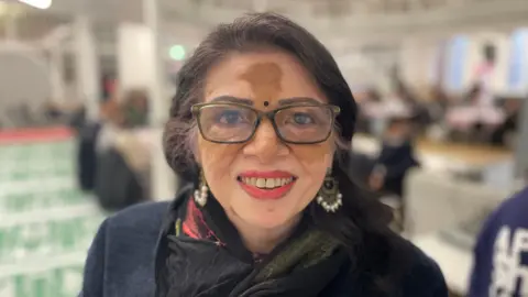 Rita Patel smiles at the camera with people in the neighbourhood centre in the background 