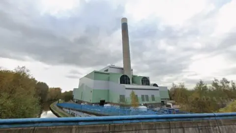 Google A factory with a tall chimney is seen at the side of a canal. It has green and grey sides and blue railings alongside it. The site is surrounded by trees.